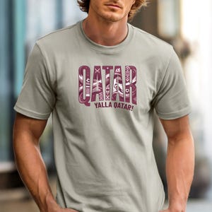 Puede incluir: Una camiseta gris claro con la palabra "QATAR" en una fuente decorativa de color granate. El texto "YALLA QATAR!" est&aacute; debajo. La camiseta es de manga corta y tiene cuello redondo.