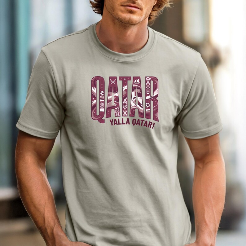 Puede incluir: Una camiseta gris claro con la palabra "QATAR" en una fuente decorativa de color granate. El texto "YALLA QATAR!" est&aacute; debajo. La camiseta es de manga corta y tiene cuello redondo.