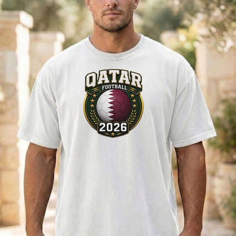 May include: White t-shirt featuring a Qatar Football emblem. The emblem includes a soccer ball with maroon and white panels, the text "QATAR FOOTBALL" in gold, and the year "2026". The design is set against a green and gold background.