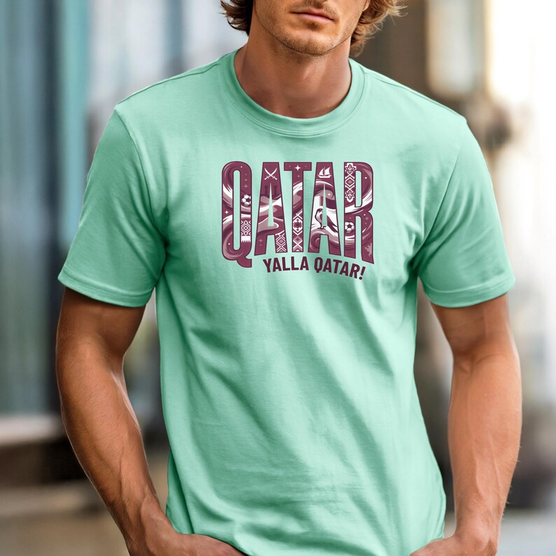 Puede incluir: Una camiseta verde claro con la palabra "QATAR" en fuente granate estampada. Debajo, la frase "YALLA QATAR!" en granate. La camiseta es de manga corta y tiene cuello redondo.