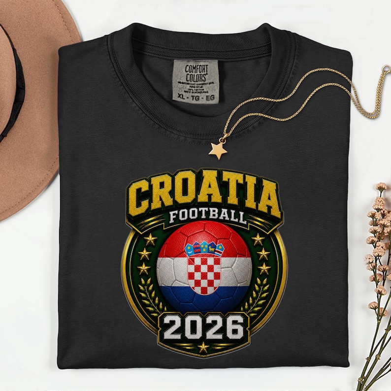 Peut inclure: T-shirt noir avec un motif de football croate. Le motif pr&eacute;sente un ballon de football avec le drapeau croate, le mot "CROATIA" en lettres dor&eacute;es et l'ann&eacute;e "2026". Un collier &eacute;toile dor&eacute; est pos&eacute; sur le t-shirt.