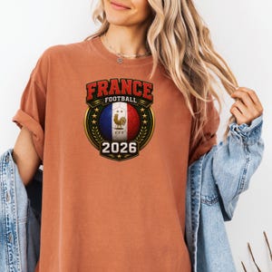 以下が含まれることがあります： フランスフットボールのグラフィックが特徴の、オレンジ色のTシャツ。フランス国旗の色を使ったサッカーボール、テキスト「FRANCE FOOTBALL」、そして年号「2026」が含まれています。