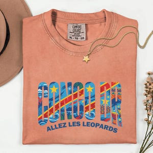 May include: Coral-colored t-shirt with the text "CONGO DR" in a colorful design, featuring the flag of the Democratic Republic of the Congo. The text "ALLEZ LES LEOPARDS" is below. A gold star pendant necklace is on top.
