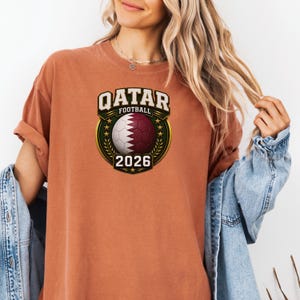 May include: A rust-colored t-shirt featuring a graphic for the Qatar Football World Cup 2026. The design includes a soccer ball with the Qatar flag colors, surrounded by a gold emblem and the text "QATAR FOOTBALL 2026".