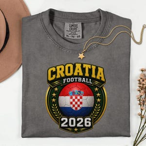 Peut inclure: Un t-shirt gris avec un motif "Croatie Football". Le motif pr&eacute;sente un ballon de football avec le drapeau croate, le mot "CROATIE" en jaune et l'ann&eacute;e "2026". Un collier dor&eacute; avec un pendentif en forme d'&eacute;toile est pos&eacute; sur le t-shirt.