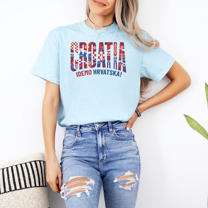 May include: Light blue t-shirt with the word "CROATIA" in a decorative, multi-colored design. Below the word is the phrase "IDEMO HRVATSKA!" in white. The shirt is worn by a person in blue jeans.