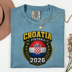 Peut inclure: Un t-shirt bleu clair avec un motif de football croate. Le motif comprend le mot "CROATIA" en lettres dor&eacute;es, un ballon de football avec le drapeau croate et l'ann&eacute;e "2026". Un collier pendentif &eacute;toile en or est &eacute;galement visible.
