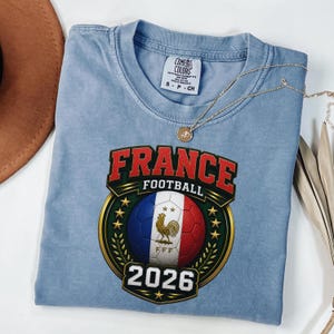 以下が含まれることがあります： 「FRANCE FOOTBALL 2026」と書かれたグラフィックが特徴の、水色のTシャツ。フランス国旗の色を使ったサッカーボール、雄鶏のエンブレム、金のリースがデザインされています。