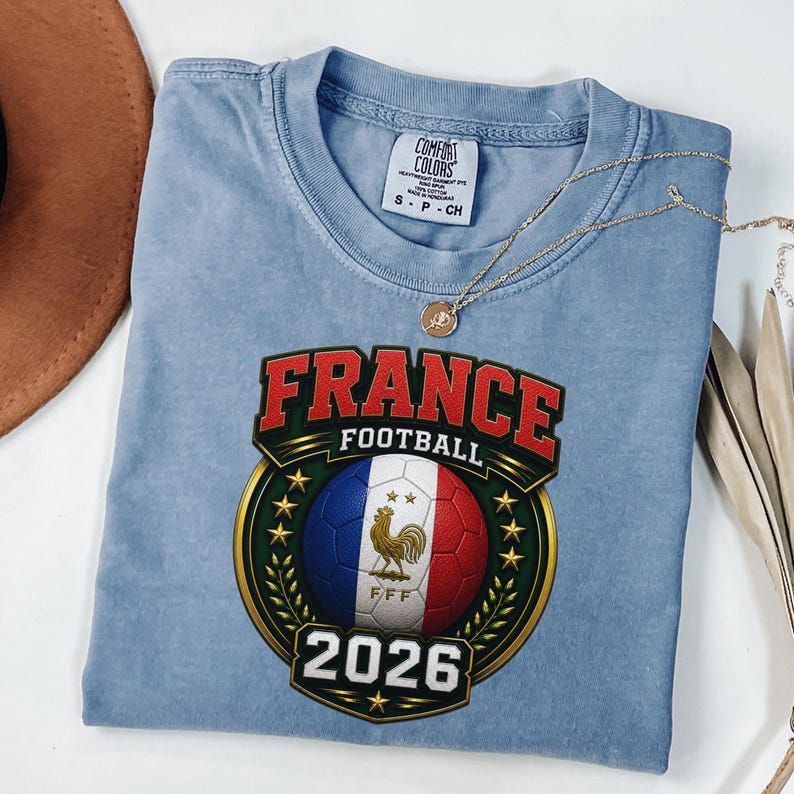 以下が含まれることがあります： 「FRANCE FOOTBALL 2026」と書かれたグラフィックが特徴の、水色のTシャツ。フランス国旗の色を使ったサッカーボール、雄鶏のエンブレム、金のリースがデザインされています。