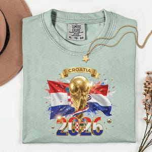 May include: A light green t-shirt featuring a graphic of the Croatian flag, a golden trophy, and the text "CROATIA 2026". A gold necklace with a star pendant rests on the shirt.