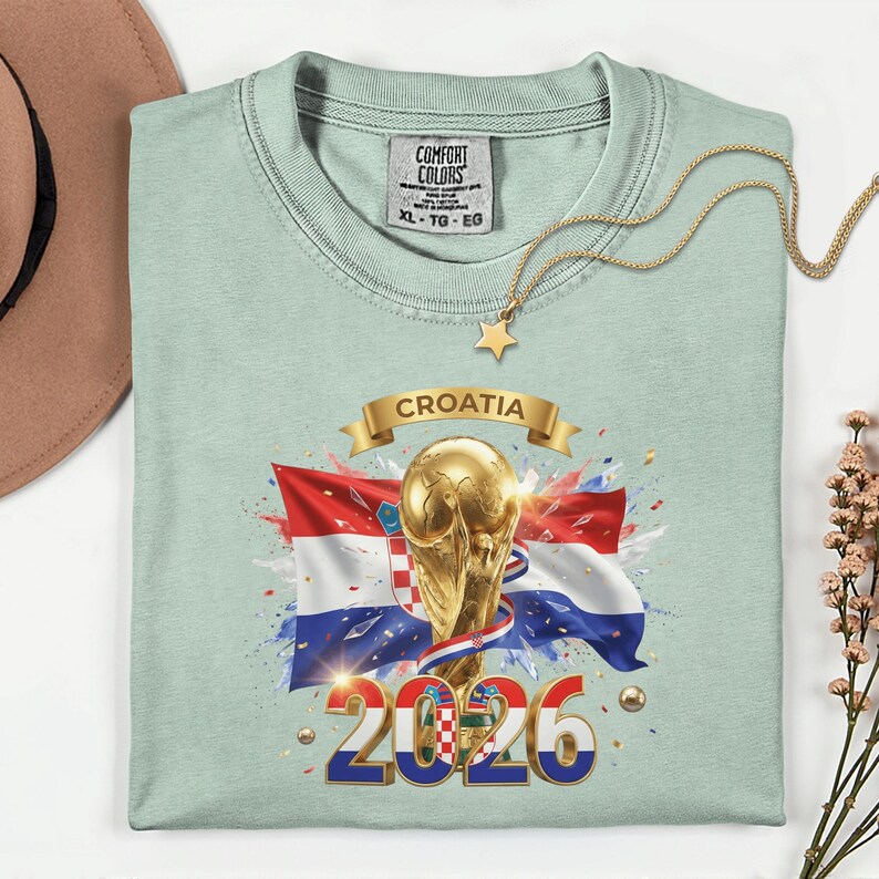 May include: A light green t-shirt featuring a graphic of the Croatian flag, a golden trophy, and the text "CROATIA 2026". A gold necklace with a star pendant rests on the shirt.