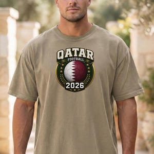 May include: A khaki-colored t-shirt featuring a Qatar Football emblem. The emblem includes a soccer ball with white and maroon panels, the text "QATAR FOOTBALL" above, and "2026" below. The design is framed by a gold wreath.