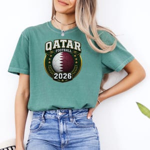 May include: A teal-colored t-shirt featuring a graphic design for Qatar Football 2026. The design includes the text "QATAR FOOTBALL" above a soccer ball with the Qatar flag colors, and the year "2026" below. The shirt is worn by a person.