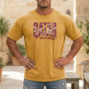 Puede incluir: Camiseta de color amarillo mostaza con la palabra "QATAR" en un dise&ntilde;o granate estampado. Debajo, la frase "YALLA QATAR!" en blanco. La camiseta tiene mangas cortas y cuello redondo.