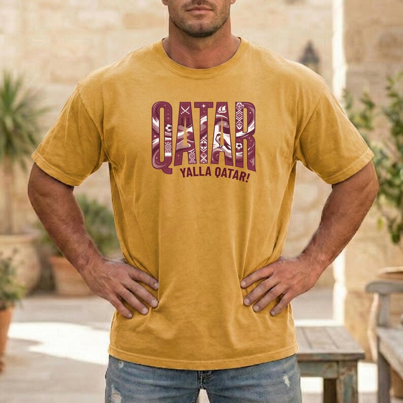 Puede incluir: Camiseta de color amarillo mostaza con la palabra "QATAR" en un dise&ntilde;o granate estampado. Debajo, la frase "YALLA QATAR!" en blanco. La camiseta tiene mangas cortas y cuello redondo.