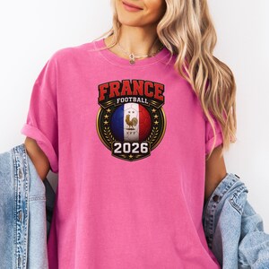 以下が含まれることがあります： フランスの国旗の色と雄鶏が描かれたサッカーボールのグラフィックが特徴のピンク色のTシャツ。「FRANCE FOOTBALL」と「2026」の文字も表示されています。カジュアルなスタイルです。