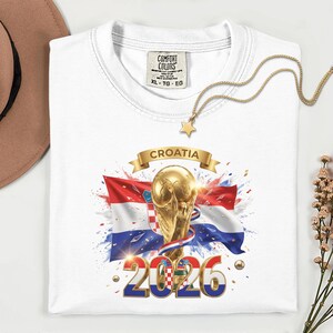 May include: White t-shirt with a graphic featuring the Croatian flag, a golden trophy, and the text "CROATIA 2026". The shirt has a crew neck and a tag that reads "COMFORT COLORS". A gold necklace with a star pendant is draped on the shirt.
