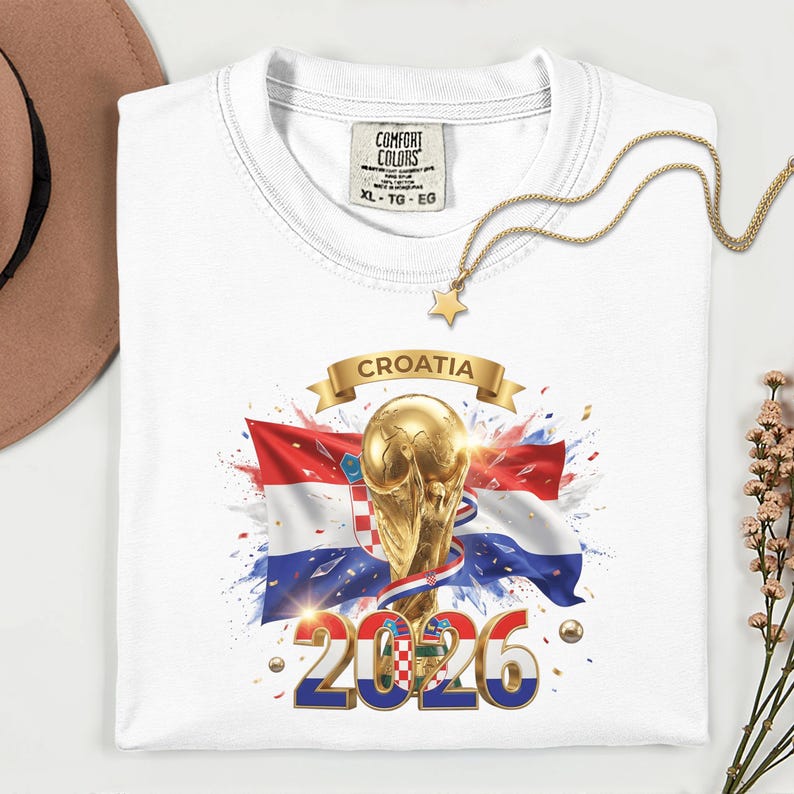 May include: White t-shirt with a graphic featuring the Croatian flag, a golden trophy, and the text "CROATIA 2026". The shirt has a crew neck and a tag that reads "COMFORT COLORS". A gold necklace with a star pendant is draped on the shirt.