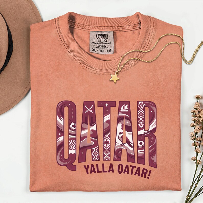 Puede incluir: Camiseta de color coral con la palabra "QATAR" en un dise&ntilde;o granate y blanco, y el texto "YALLA QATAR!". Se ve un collar con colgante de estrella dorada y un sombrero.