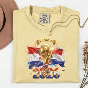 May include: A light yellow t-shirt featuring a graphic of the Croatian flag, a golden trophy, and the text "CROATIA 2026". The shirt has a crew neck and short sleeves. A gold necklace with a star pendant is draped across the shirt.
