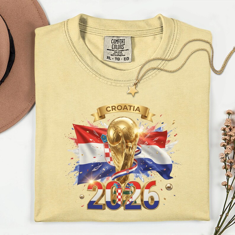May include: A light yellow t-shirt featuring a graphic of the Croatian flag, a golden trophy, and the text "CROATIA 2026". The shirt has a crew neck and short sleeves. A gold necklace with a star pendant is draped across the shirt.