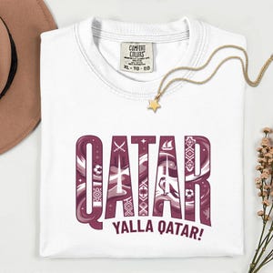 Puede incluir: Camiseta blanca con la palabra "QATAR" en un dise&ntilde;o granate y blanco, con el texto "YALLA QATAR!". La camiseta tiene una etiqueta Comfort Colors y se combina con un collar dorado y un sombrero marr&oacute;n.