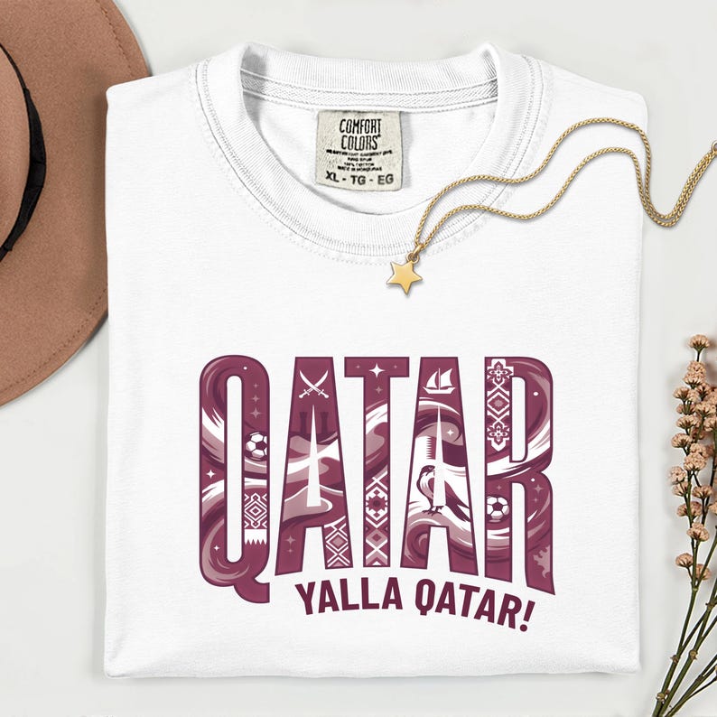Puede incluir: Camiseta blanca con la palabra "QATAR" en un dise&ntilde;o granate y blanco, con el texto "YALLA QATAR!". La camiseta tiene una etiqueta Comfort Colors y se combina con un collar dorado y un sombrero marr&oacute;n.
