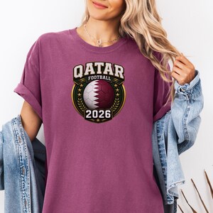May include: A maroon t-shirt featuring a soccer ball with the Qatar flag design, the text "QATAR FOOTBALL" above, and "2026" below. The design is in a gold and green circular emblem.