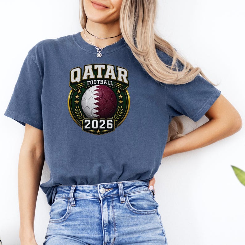 May include: A blue t-shirt featuring a Qatar Football design. The design includes a soccer ball with the Qatar flag colors, the text "QATAR FOOTBALL 2026" in a gold and green emblem.