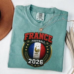 以下が含まれることがあります： 「FRANCE FOOTBALL 2026」と書かれたグラフィックが特徴の、シーフォームグリーンのTシャツ。フランス国旗の色と金の雄鶏が描かれたサッカーボールのグラフィック。ペンダント付きのシルバーネックレスがシャツの上にあります。