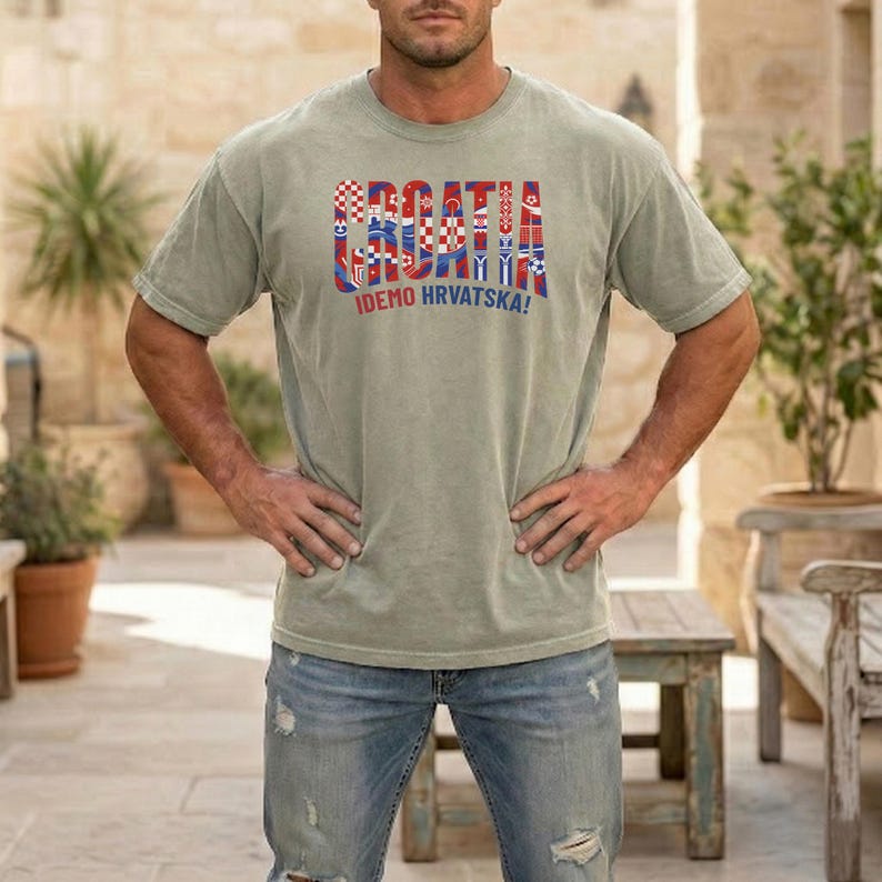 May include: A sage green t-shirt with the word "CROATIA" in a multi-colored, flag-inspired design. Below the word "CROATIA" is the text "IDEMO HRVATSKA!" in blue and red. The shirt is made of a soft material.