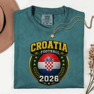 Peut inclure: T-shirt de couleur sarcelle avec un &eacute;cusson dor&eacute; affichant "CROATIA FOOTBALL" et un ballon de football aux couleurs du drapeau croate. L'ann&eacute;e "2026" est en dessous. Un collier pendentif &eacute;toile dor&eacute;e est pos&eacute; sur le t-shirt.
