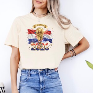 May include: Cream-colored t-shirt featuring a graphic of the Croatian flag, a golden trophy, and the year "2026." The word "Croatia" is displayed on a banner above the trophy. The design includes red, white, and blue colors.