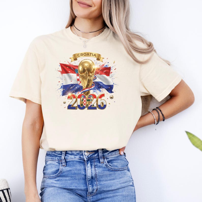 May include: Cream-colored t-shirt featuring a graphic of the Croatian flag, a golden trophy, and the year "2026." The word "Croatia" is displayed on a banner above the trophy. The design includes red, white, and blue colors.