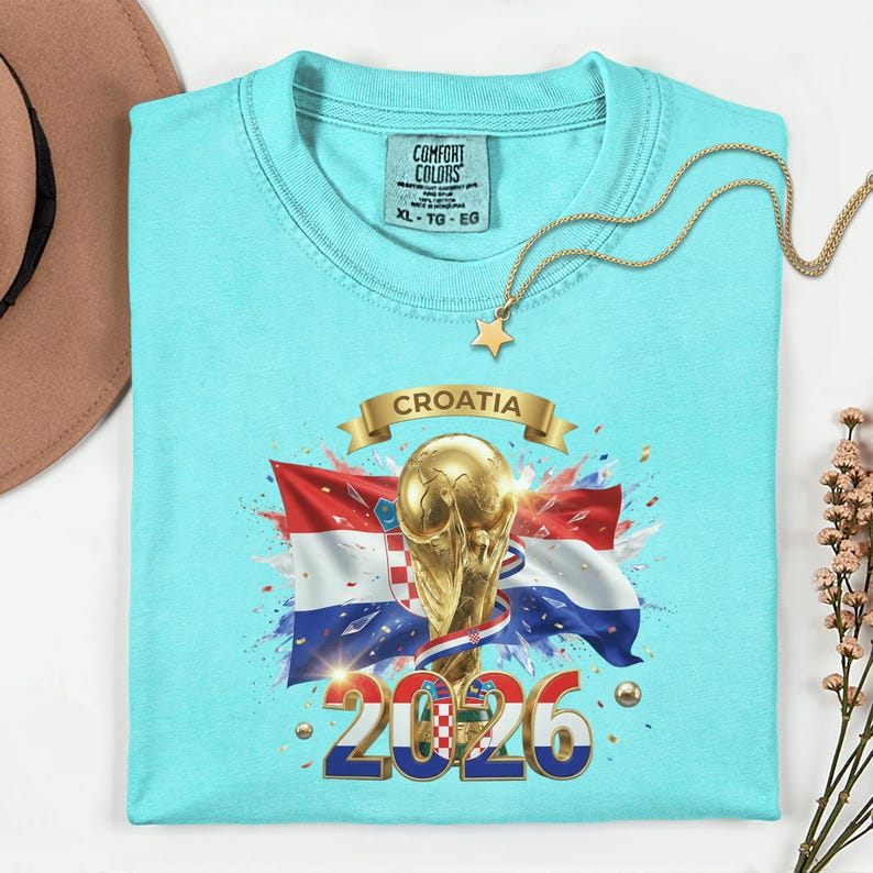 May include: A light blue t-shirt featuring a graphic of the Croatian flag, a golden trophy, and the text "CROATIA 2026." The shirt has a crew neck and a tag that reads "Comfort Colors XL-TG-EG."