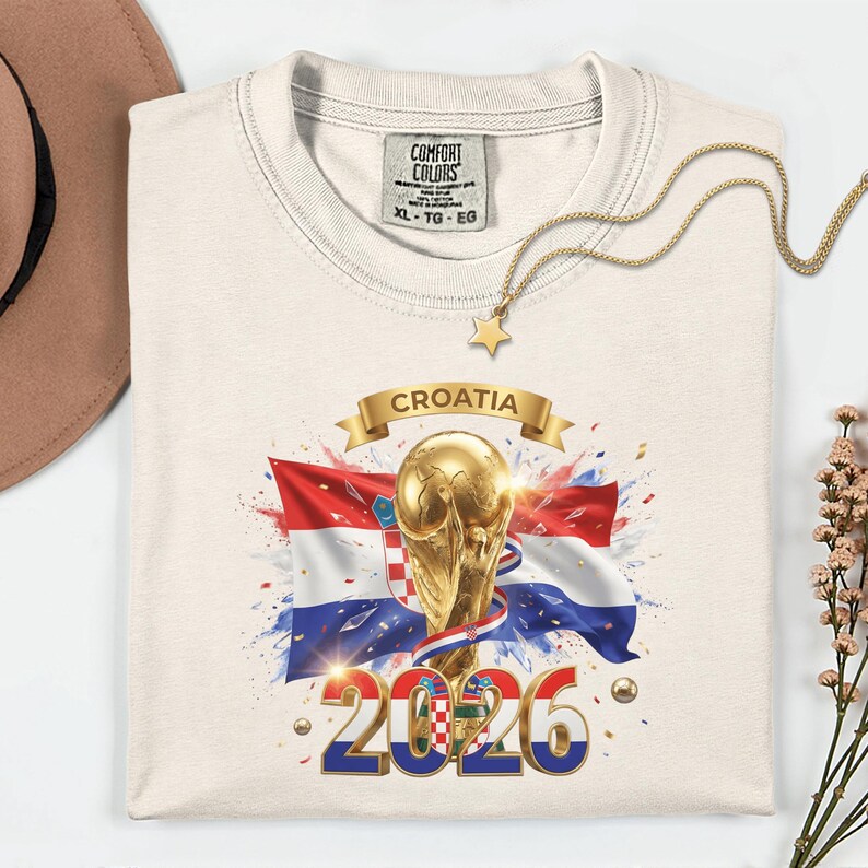 May include: Cream-colored t-shirt featuring a Croatian flag design with a golden trophy, the word "CROATIA" on a banner, and the year "2026". The shirt has a Comfort Colors tag and a gold star necklace.