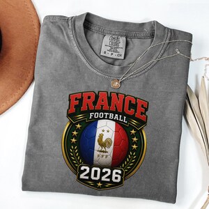 以下が含まれることがあります： フランス国旗の色をしたサッカーボール、金の雄鶏、そして「FRANCE FOOTBALL 2026」の文字が描かれたグレーのComfort Colors Tシャツ。 ペンダント付きのシルバーネックレスがシャツの上にかけられています。