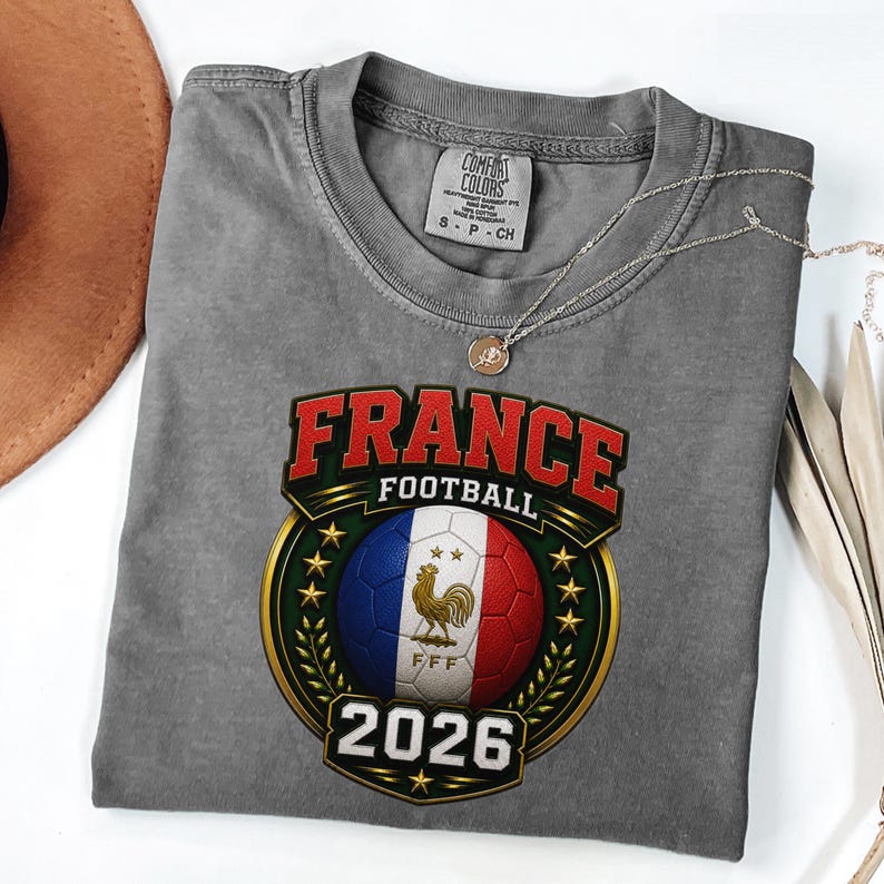 以下が含まれることがあります： フランス国旗の色をしたサッカーボール、金の雄鶏、そして「FRANCE FOOTBALL 2026」の文字が描かれたグレーのComfort Colors Tシャツ。 ペンダント付きのシルバーネックレスがシャツの上にかけられています。