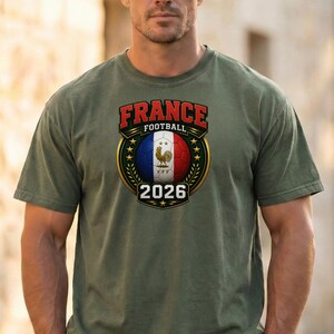 以下が含まれることがあります： オリーブグリーンのTシャツ。フランス国旗の色、雄鶏、そして「FRANCE FOOTBALL 2026」の文字が入ったサッカーボールのグラフィック。デザインは金の縁と星で囲まれています。