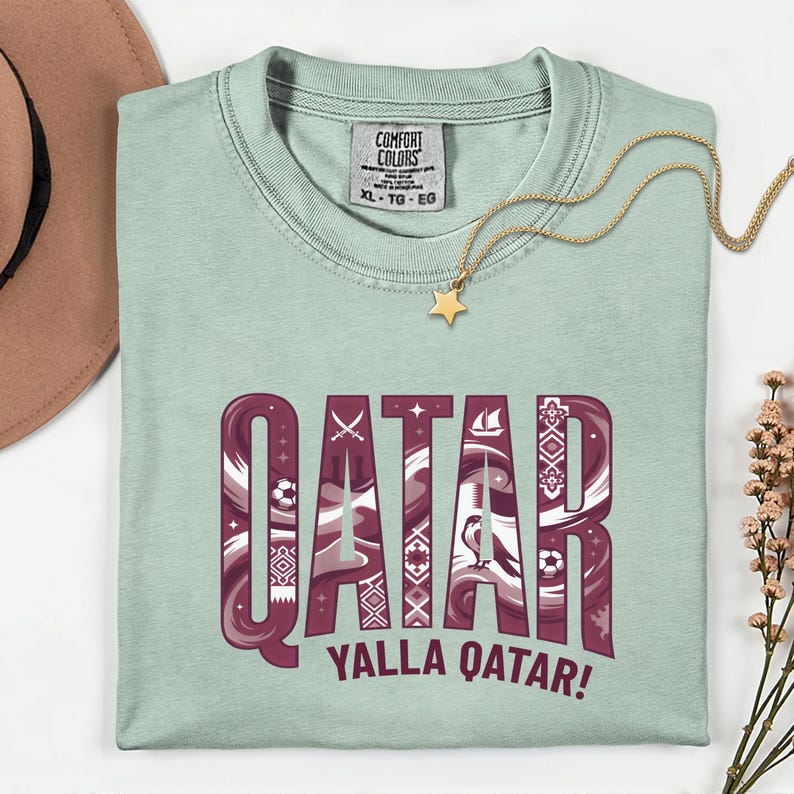 Puede incluir: Camiseta verde claro con la palabra "QATAR" en un dise&ntilde;o granate estampado. El texto "YALLA QATAR!" est&aacute; debajo. Se ve un collar dorado con un colgante de estrella y una etiqueta Comfort Colors.