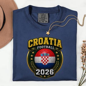 Peut inclure: T-shirt bleu marine avec un motif dor&eacute; et vert repr&eacute;sentant le mot "CROATIA FOOTBALL" au-dessus d'un ballon de football aux couleurs du drapeau croate. L'ann&eacute;e "2026" est en dessous. Un collier &eacute;toile dor&eacute; est pos&eacute; sur le t-shirt.