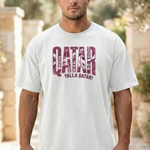 Puede incluir: Camiseta blanca con un gr&aacute;fico granate que dice "QATAR" en una fuente estilizada, con elementos decorativos. Debajo de la palabra "QATAR" est&aacute; la frase "YALLA QATAR!" en granate. La camiseta tiene mangas cortas y cuello redondo.