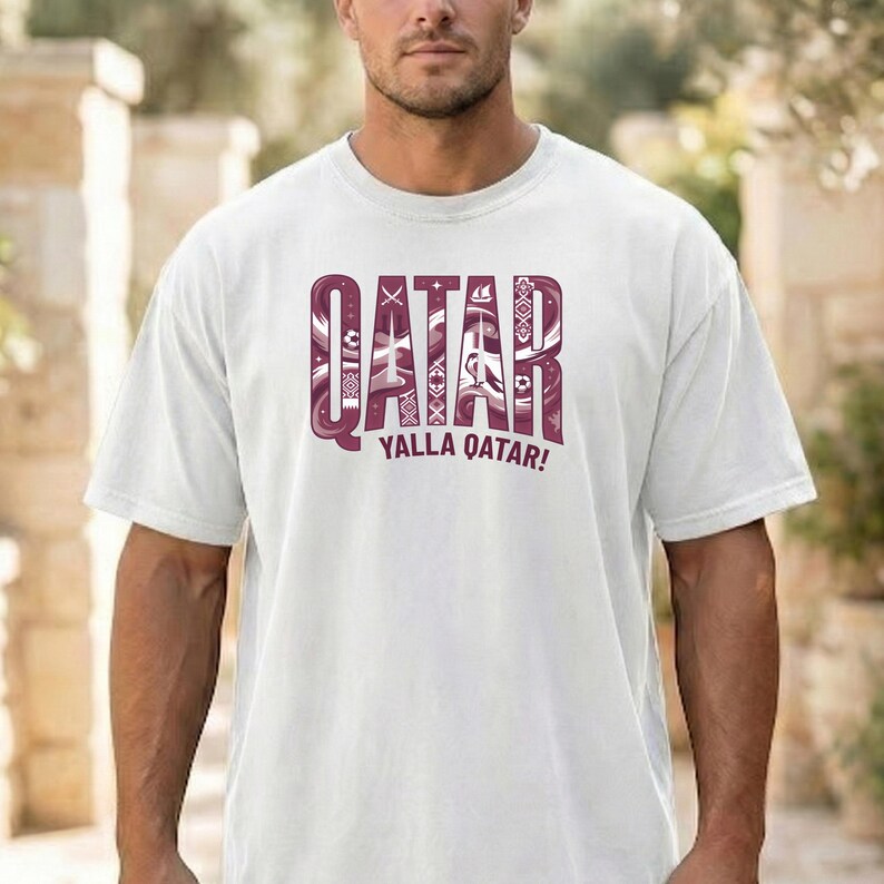 Puede incluir: Camiseta blanca con un gr&aacute;fico granate que dice "QATAR" en una fuente estilizada, con elementos decorativos. Debajo de la palabra "QATAR" est&aacute; la frase "YALLA QATAR!" en granate. La camiseta tiene mangas cortas y cuello redondo.