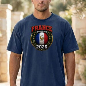 以下が含まれることがあります： フランス国旗の色と雄鶏のエンブレムが付いたサッカーボールのグラフィックが特徴のネイビーブルーのTシャツ。テキスト「FRANCE FOOTBALL 2026」がグラフィックの上部と下部に表示されています。柔らかい素材で作られたシャツです。