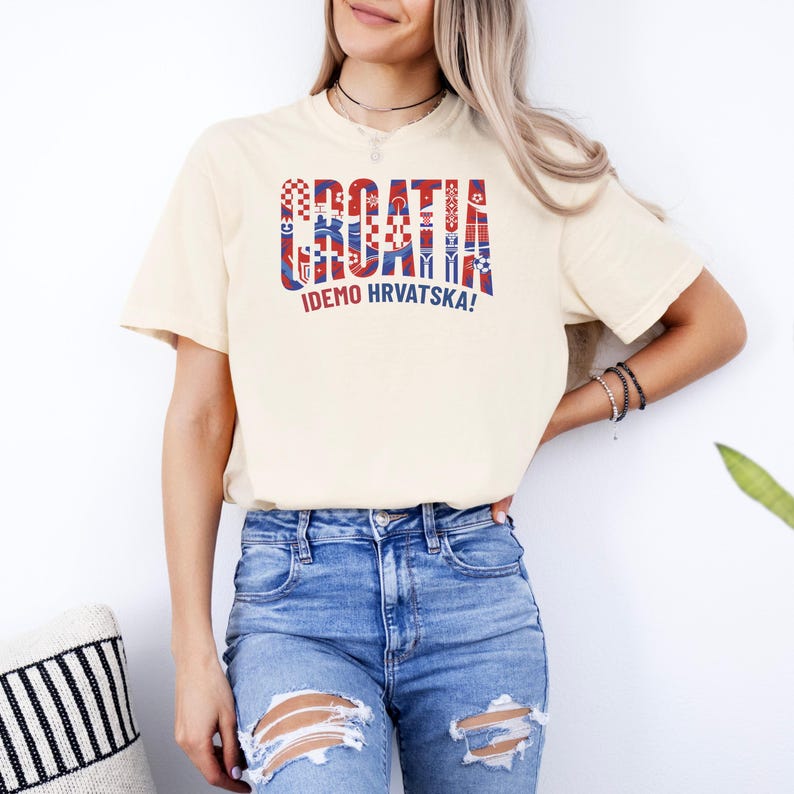 May include: Cream-colored t-shirt with the word "CROATIA" in red and blue, with decorative patterns. Below the word is the phrase "IDEMO HRVATSKA!" in green. The shirt is paired with ripped blue jeans.