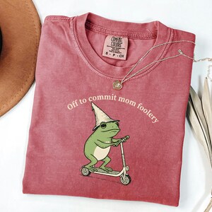 May include: A red Comfort Colors t-shirt with the phrase "Off to commit mom foolery" and a graphic of a green frog riding a scooter. A gold necklace is draped across the shirt.