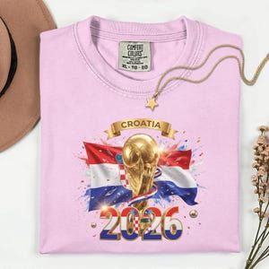 May include: A light pink t-shirt featuring a graphic of the Croatian flag, a golden trophy, and the text "CROATIA 2026." The shirt has a crew neck and short sleeves. A gold necklace with a star pendant is draped on the shirt.