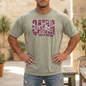 Puede incluir: Una camiseta verde claro con la palabra "QATAR" en una fuente granate decorativa. Debajo de la palabra "QATAR" est&aacute; la frase "YALLA QATAR!" en granate. La camiseta la lleva una persona.