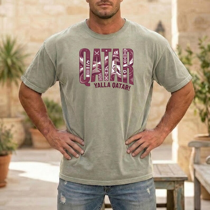 Puede incluir: Una camiseta verde claro con la palabra "QATAR" en una fuente granate decorativa. Debajo de la palabra "QATAR" est&aacute; la frase "YALLA QATAR!" en granate. La camiseta la lleva una persona.
