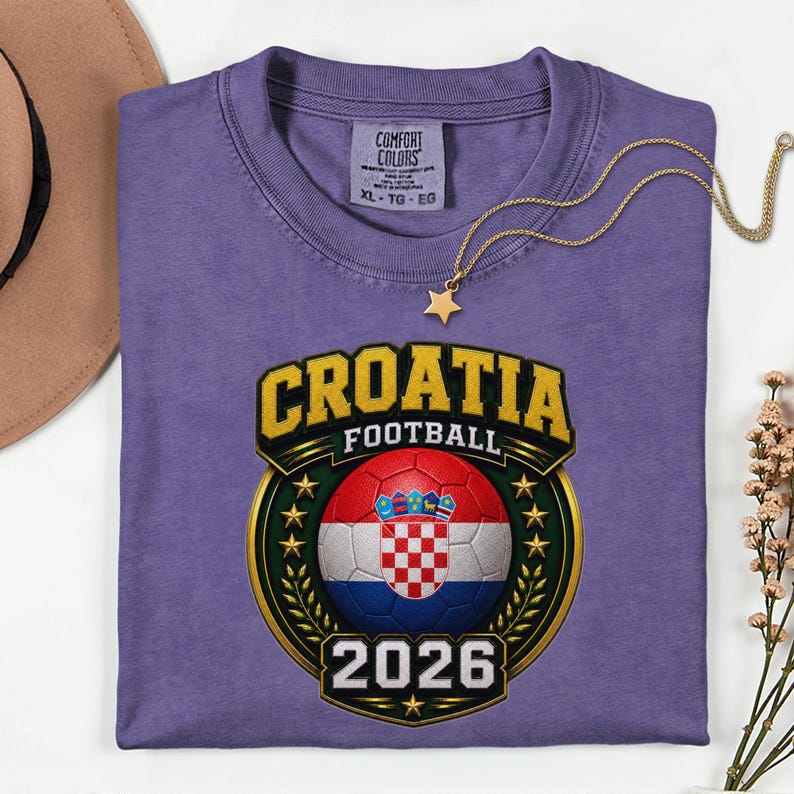 Peut inclure: Un t-shirt violet avec un motif dor&eacute; et vert indiquant "CROATIE FOOTBALL 2026" avec un ballon de football aux couleurs du drapeau croate. Un collier dor&eacute; avec un pendentif en forme d'&eacute;toile est pos&eacute; sur le t-shirt.